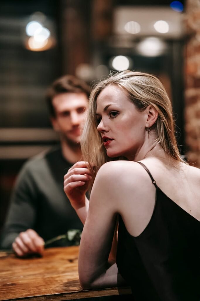 Dreamy girlfriend in black dress looking over shoulder while sitting at wooden table with unrecognizable boyfriend on blurred background during romantic date