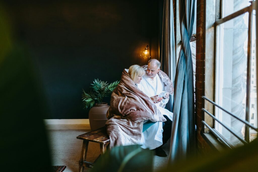 A senior couple wrapped in a blanket, sharing a warm moment by the window indoors.